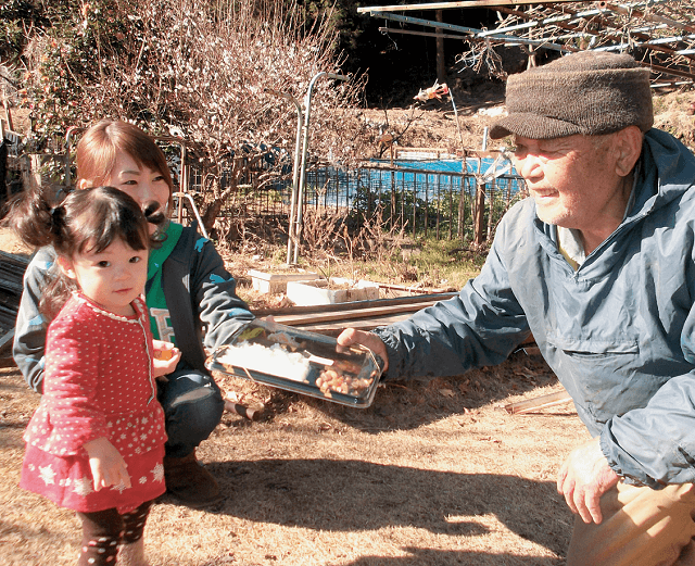 写真2：かわね来風が行う「ママ宅事業」の様子を表す写真。幼児とママさんが高齢者宅を訪れお弁当を届ける高齢者宅配サービス事業。